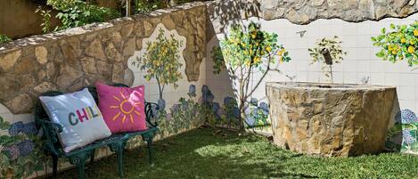 Un muro de piedra con un mural de azulejos, un banco con cojines y un tocón de árbol.