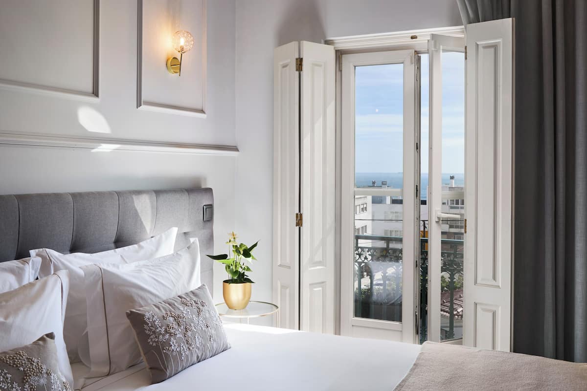 A hotel room with a bed, a potted plant, a wall-mounted light fixture, and a view through an open door.