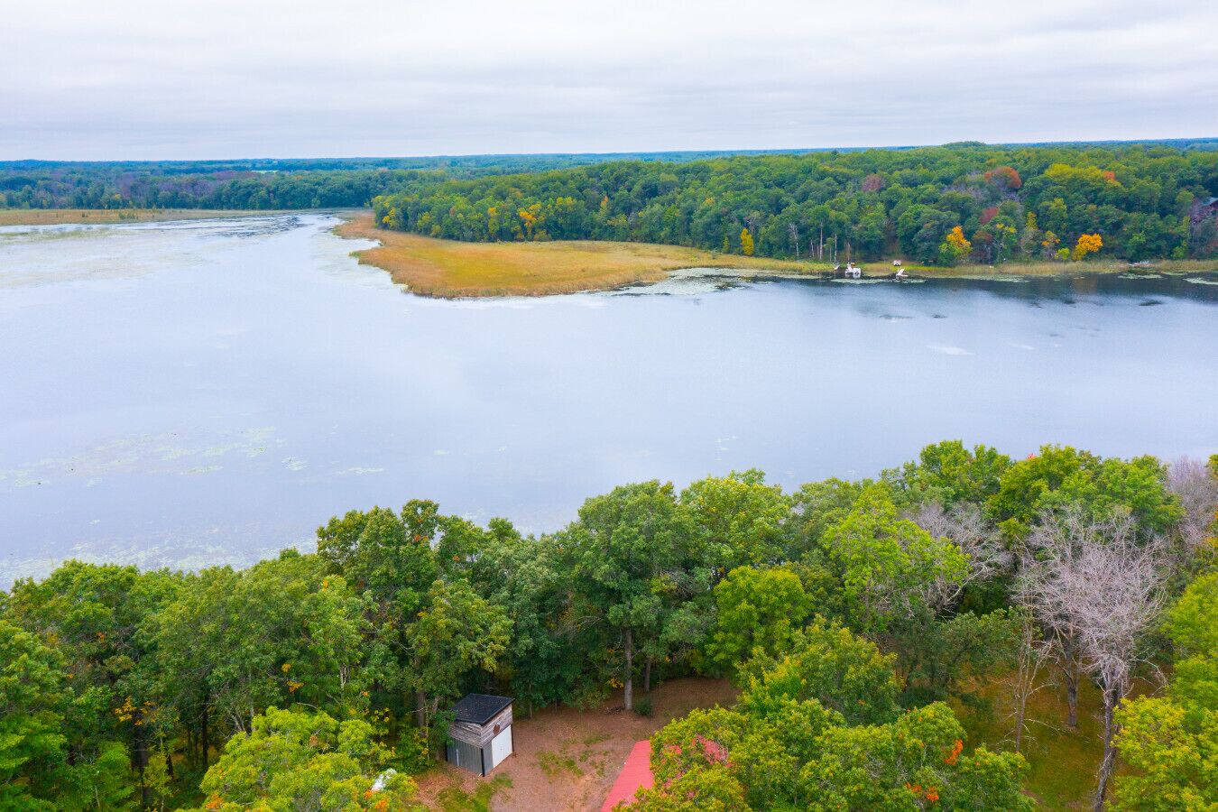 Una propiedad junto al lago con un bosque denso, una cabaña pequeña y un sendero rojo.