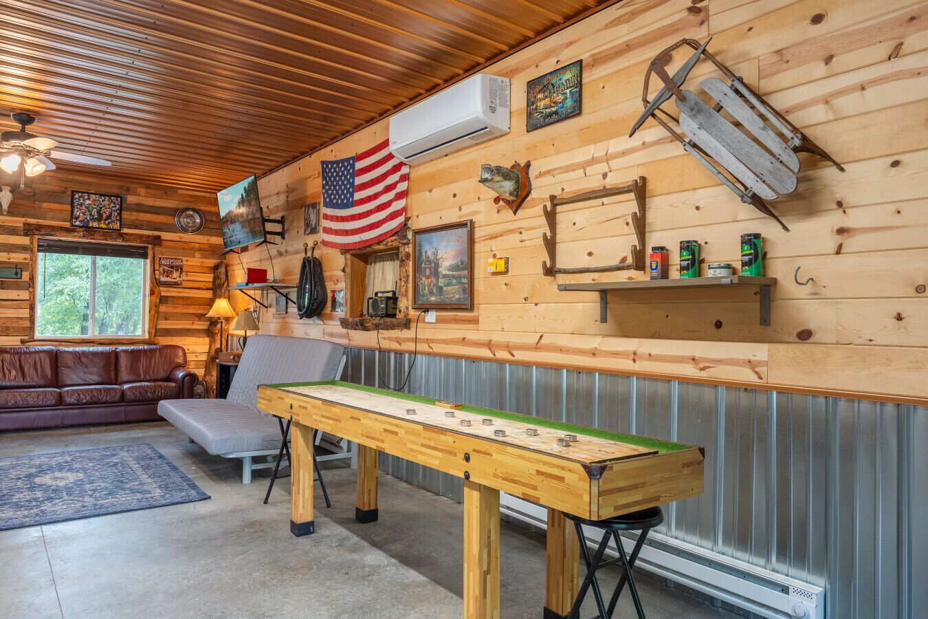A cozy cabin interior with a wooden ceiling, a large American flag, a fireplace, a sofa, and a table with a game.