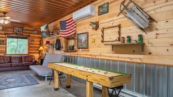 A cozy cabin interior with a wooden ceiling, a large American flag, a fireplace, a sofa, and a table with a game.