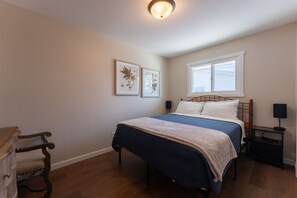 A bedroom with a bed, a nightstand, a lamp, a chair, and two framed pictures on the wall. - UWEC Campus & Clinic Retreat (Eau Claire)