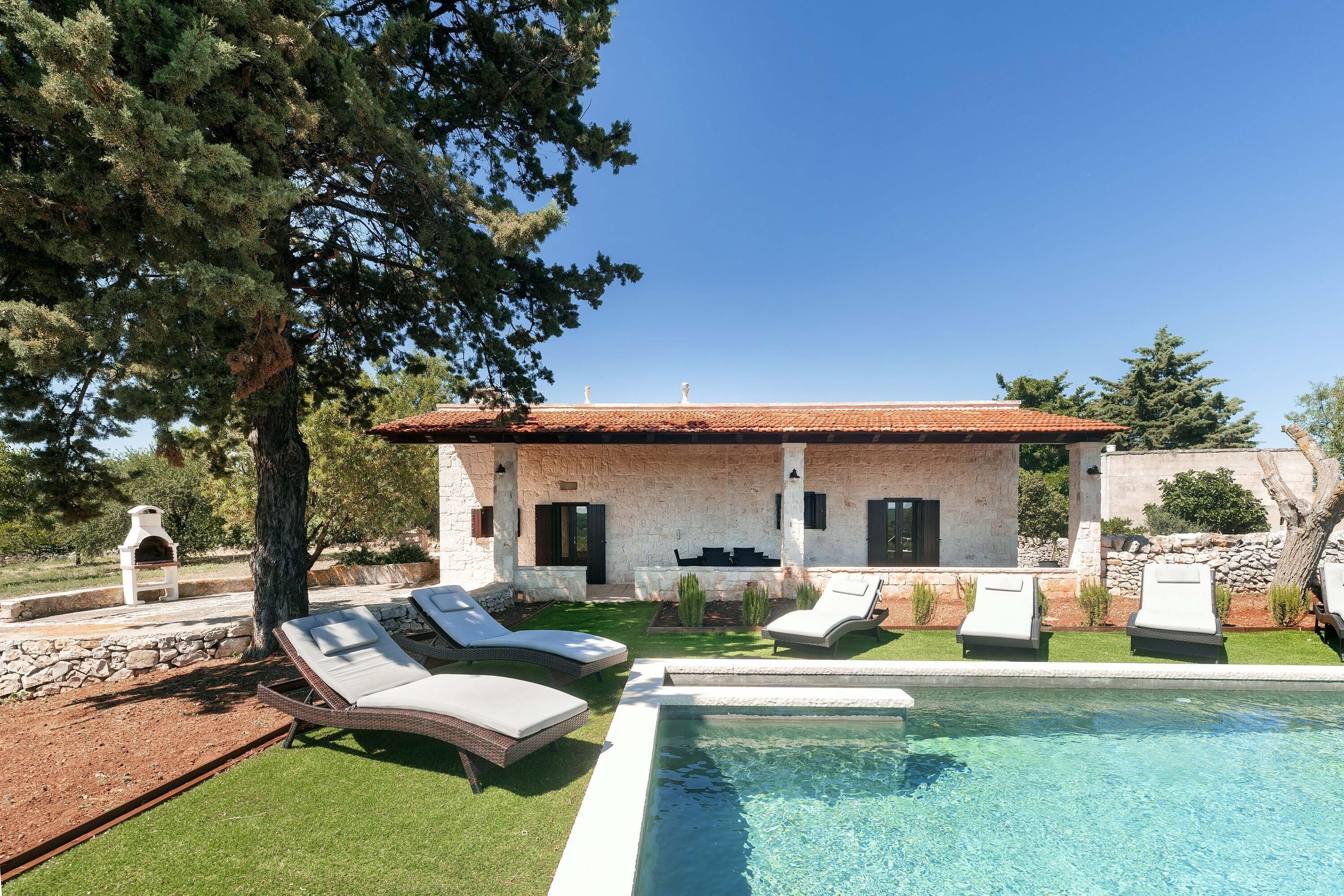 Une maison avec une piscine, des chaises longues pour l’extérieur et un mur en pierre.