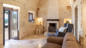 Una habitación con paredes de piedra, chimenea, un sofá, una mesita y vistas a través de un ventanal.