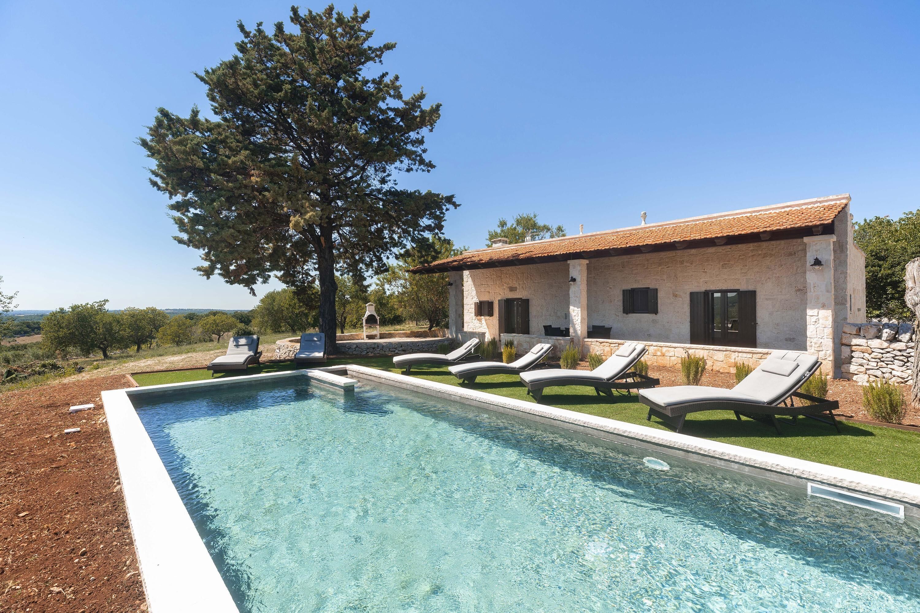 Une piscine avec des chaises longues, un bâtiment en pierre et un grand arbre.