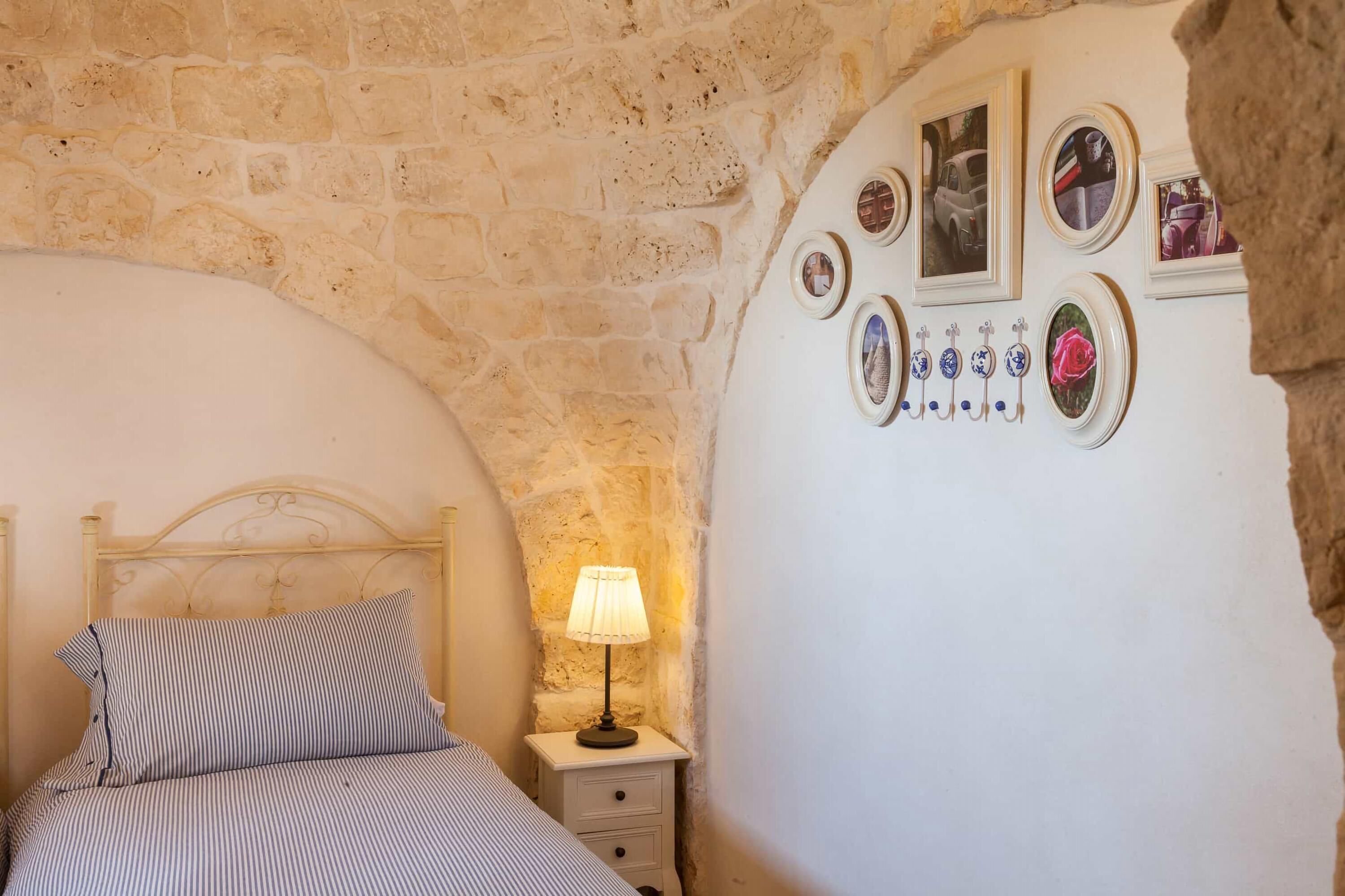 A bedroom with a stone wall, a bed with striped bedding, a bedside table with a lamp, and a wall decorated with framed pictures and a vase.