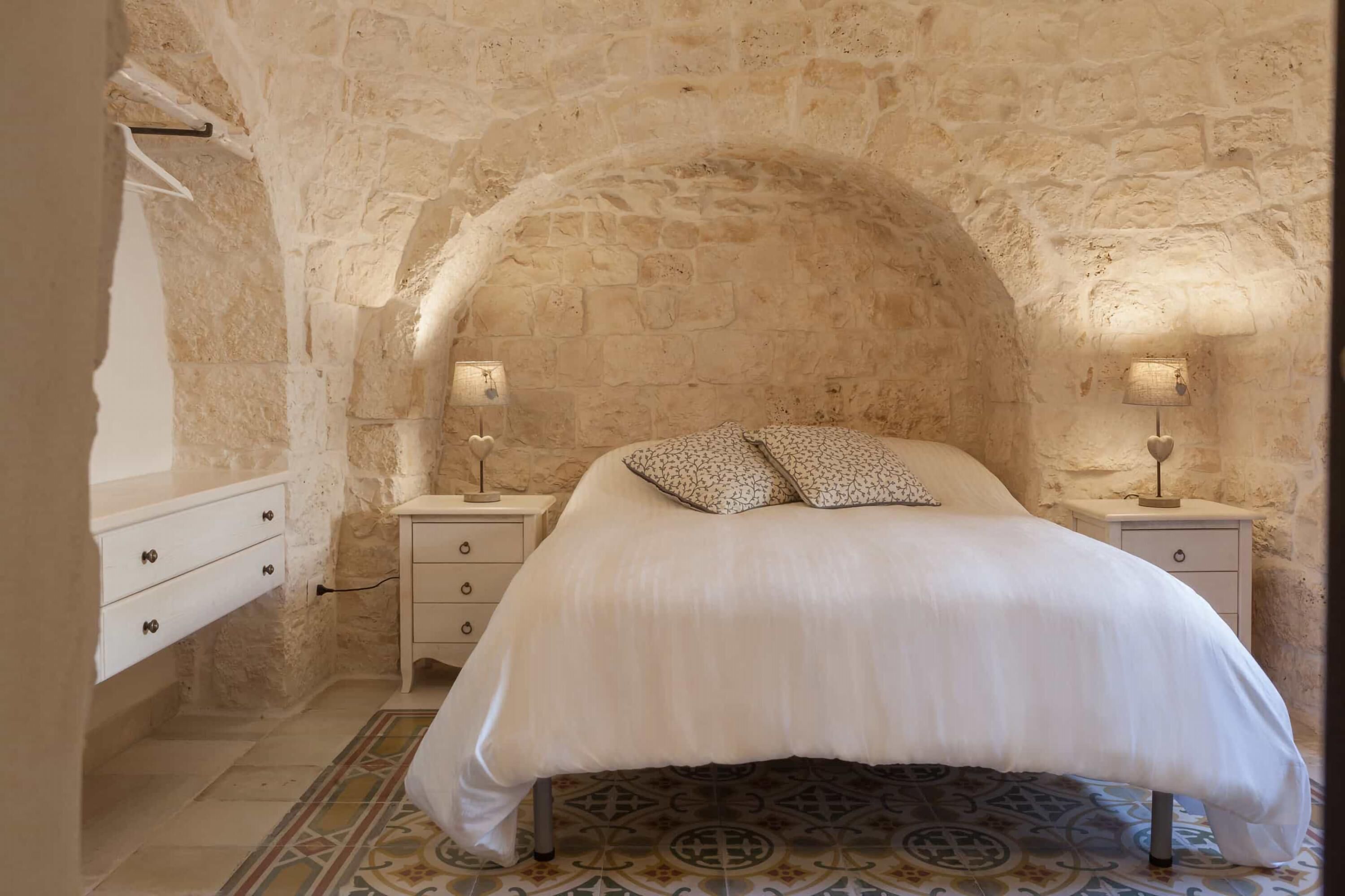 Un dormitorio con pared de piedra, cama con almohadas estampadas, mesita de noche con lámpara y suelo de baldosas.