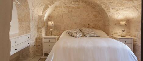 Un dormitorio con pared de piedra, cama con almohadas estampadas, mesita de noche con lámpara y suelo de baldosas.