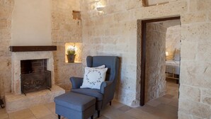 Una habitación con paredes de piedra, una chimenea, un sillón azul con un cojín decorativo, y una cama en la habitación contigua.