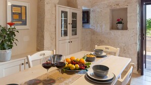 Una mesa de comedor con una copa de vino, fruta y cuencos. Hay un armario con puertas de cristal y una planta en maceta.