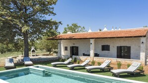 Una piscina con sillones, un edificio de piedra con techo de tejas y un árbol.