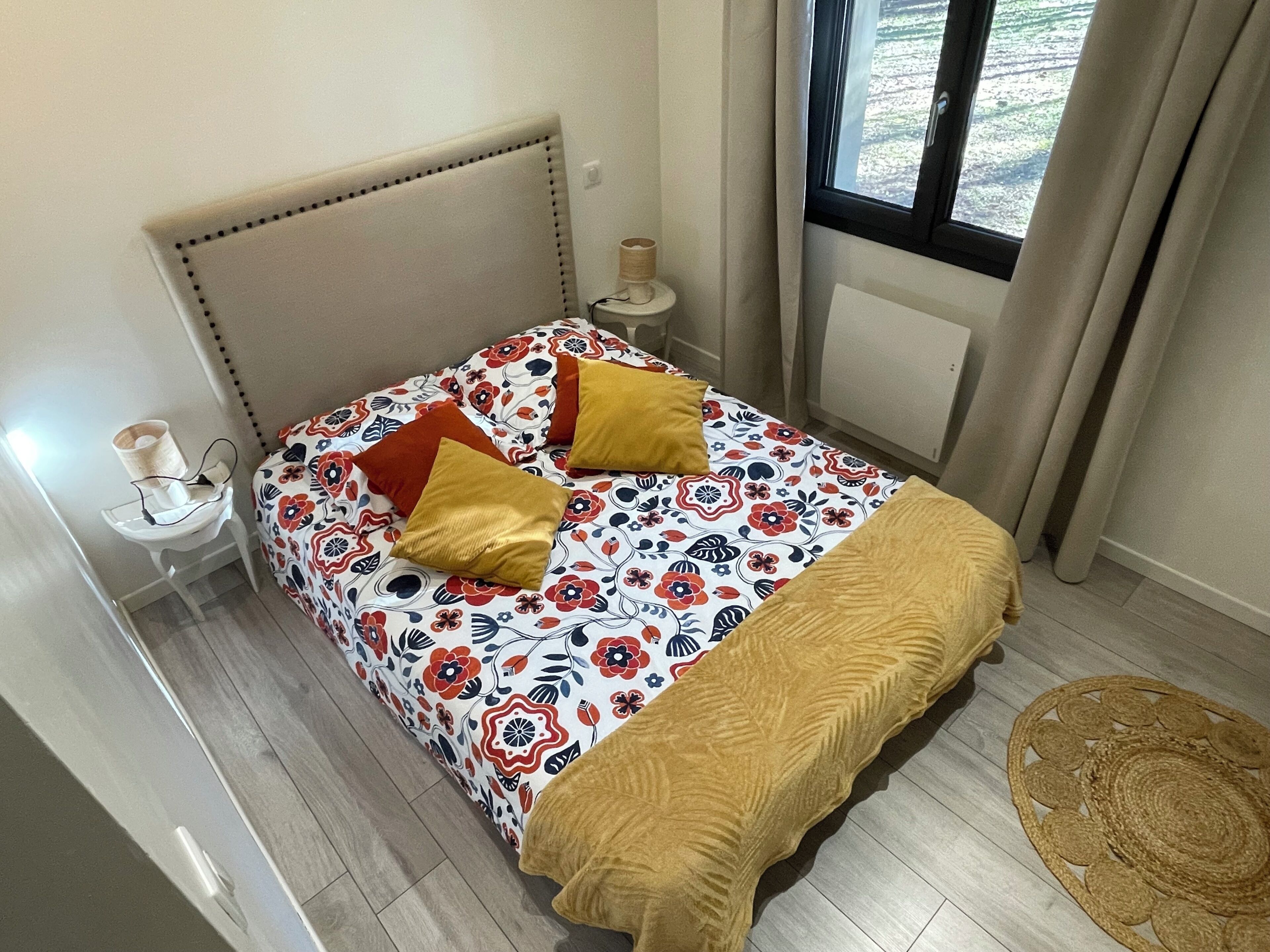 Ein Bett mit floralem Überwurf, zwei Kissen, ein Nachttisch mit Lampe und ein Fenster mit Vorhängen.