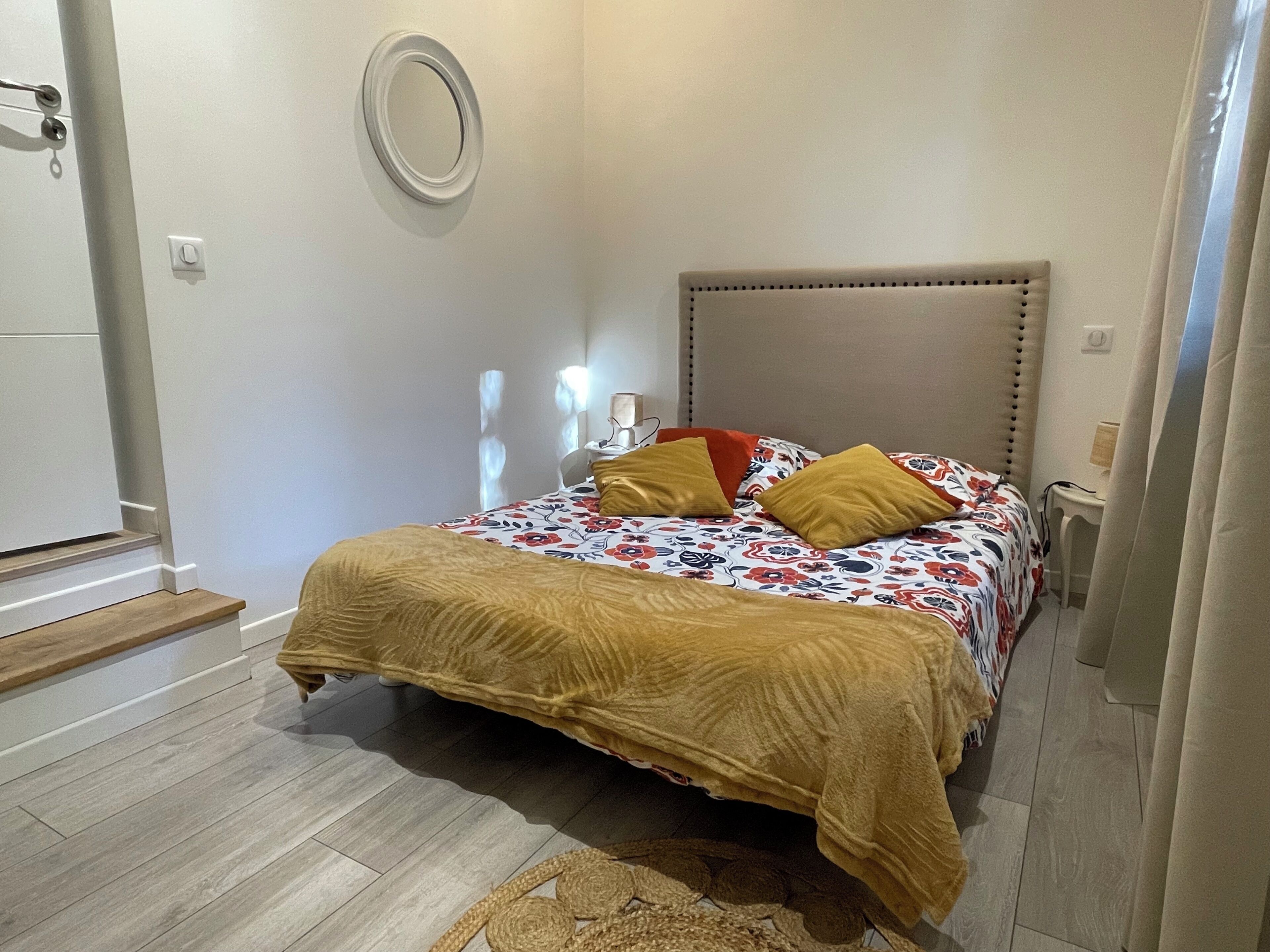 Une chambre à coucher comprenant un lit, une table de chevet, une lampe, un luminaire fixé au mur, une porte et une fenêtre avec des rideaux.