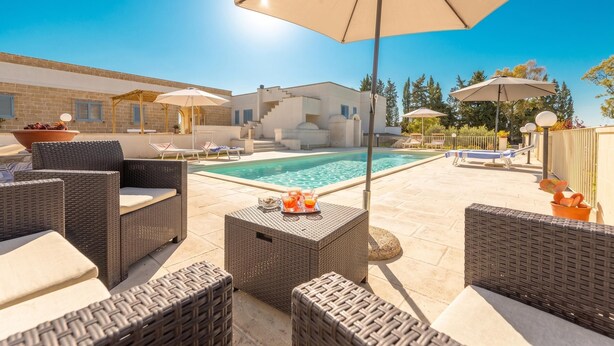 Un área junto a la piscina con muebles de mimbre, sombrillas y un cielo azul despejado.