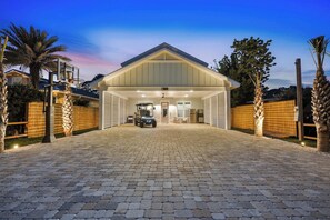 Une allée couverte mène à une maison dotée d’un garage, bordée de palmiers, sous un ciel dégagé.