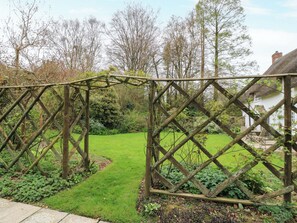 Ein hölzernes Tor mit Gitterwerk, ein grünes Rasenstück und im Hintergrund ein weißes Gebäude mit einem Reetdach.