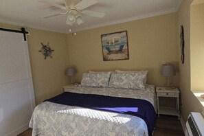 Une chambre à coucher avec un lit, un ventilateur de plafond, une table de chevet avec une lampe, une horloge murale et un tableau représentant une scène de plage.