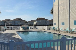 Une piscine entourée d’une clôture blanche, avec des chaises longues et un bâtiment doté de balcons.