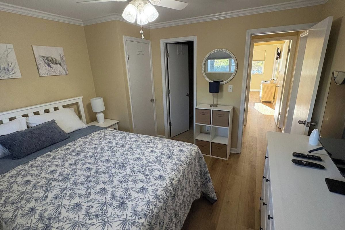 A bedroom with a bed, bedside tables, a desk with a computer, a mirror, and a door leading to a bathroom.