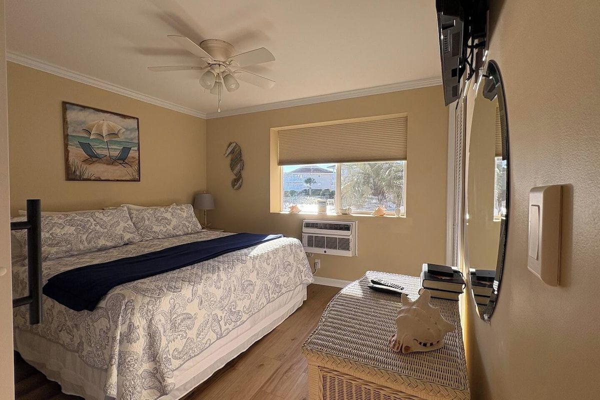 A bedroom with a bed, a ceiling fan, a window with blinds, a wall-mounted air conditioner, and a decorative item on a table.