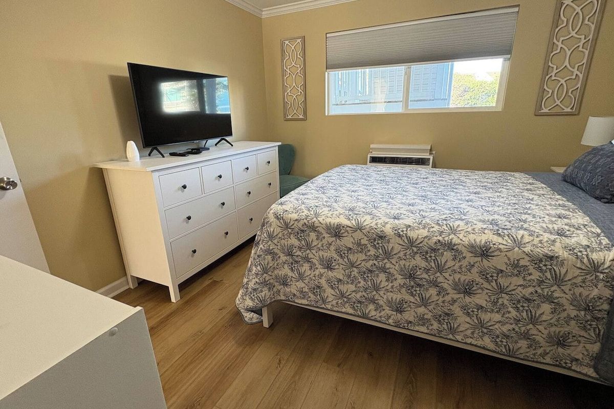 A bedroom with a bed, a television on a dresser, a window with blinds, and a decorative wall panel.