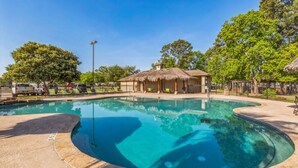 A swimming pool with a curved edge, surrounded by a fence, and a building with a thatched roof in the background.