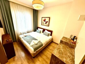 A hotel room with a bed, a bedside table, a nightstand, a marble side table, a painting, and a window with curtains.