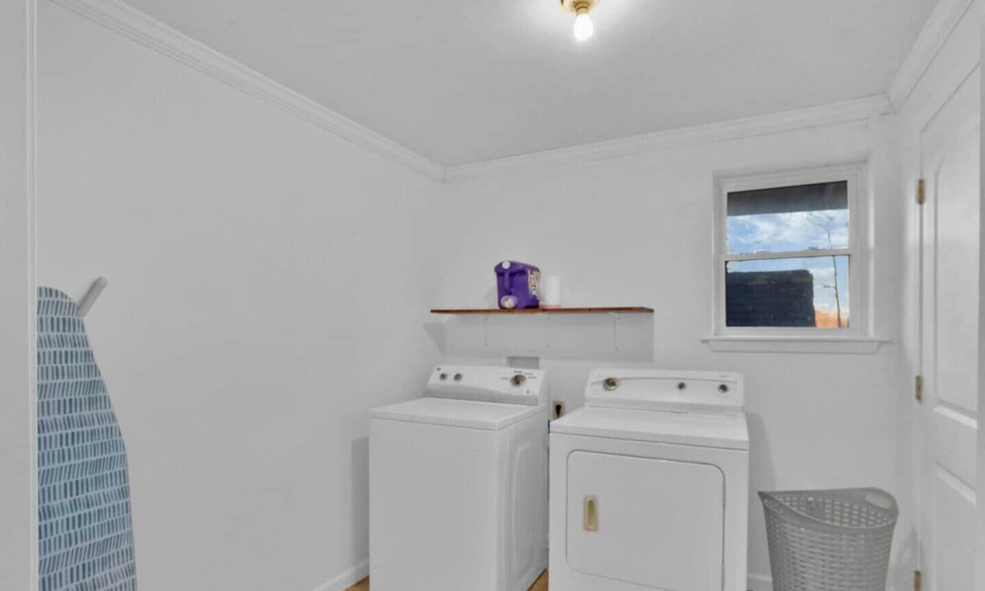 A laundry room with two white washing machines, a purple detergent box on a shelf, a window with a view, and a laundry basket.