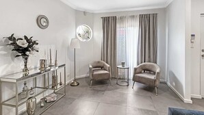 Ein stilvolles Wohnzimmer mit einem Glastische, zwei Sesseln, einem runden Tisch und einem großen Fenster mit Vorhängen.