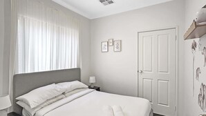 Ein ordentlich gemachtes Bett mit weißen Bettwäsche und zwei gerollten Handtüchern, ein Nachttisch mit Lampe, ein Fenster mit durchsichtigen Vorhängen und eine Tür mit Wandsticker.