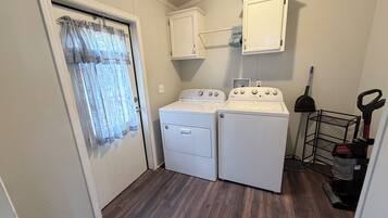A laundry room with two white washers and dryers, a metal rack, and a vacuum cleaner.