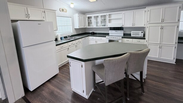 A kitchen with white cabinets, a refrigerator, a stove, and a microwave. There are two bar stools and a window with blinds. - Countryside Home in Beautiful Aiken SC (Aiken)