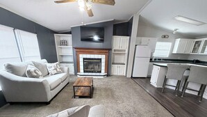 A modern living room with a fireplace, a sofa, a coffee table, and a kitchen area with white cabinets and a refrigerator. - Countryside Home in Beautiful Aiken SC (Aiken)