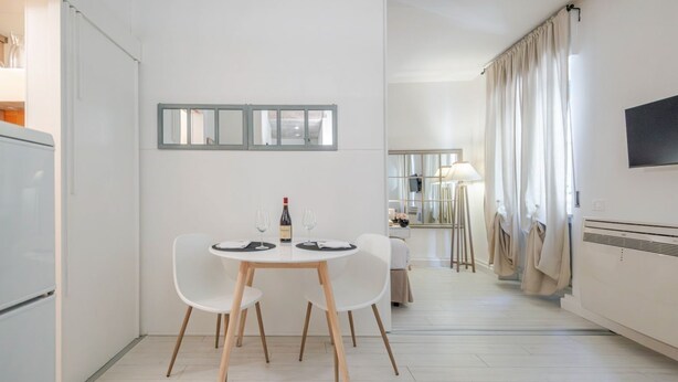 Eine moderne Küche mit weißen Schränken, ein Essbereich mit zwei weißen Stühlen, ein Tisch mit Flasche und Gläsern sowie ein an der Wand befestigter Fernseher.