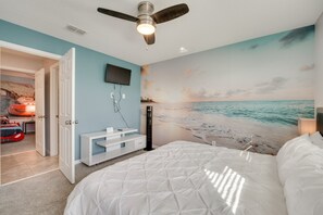 Ein Schlafzimmer mit einem Bett, einem Deckenventilator, einem an der Wand befestigten Fernseher und einem Wandgemälde mit Strandmotiv.