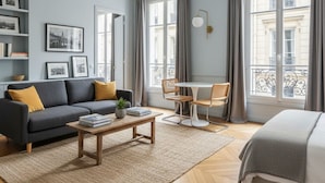 Ein modernes Wohnzimmer mit einer Couch, einem Couchtisch und einem Essbereich mit Stühlen. Große Fenster mit Vorhängen, ein Bücherregal mit gerahmten Bildern und ein Bett mit Decke.