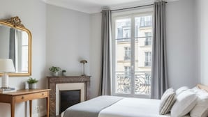 Ein Schlafzimmer mit Bett, Schreibtisch, Kamin und einem großen Fenster mit Blick auf Gebäude.