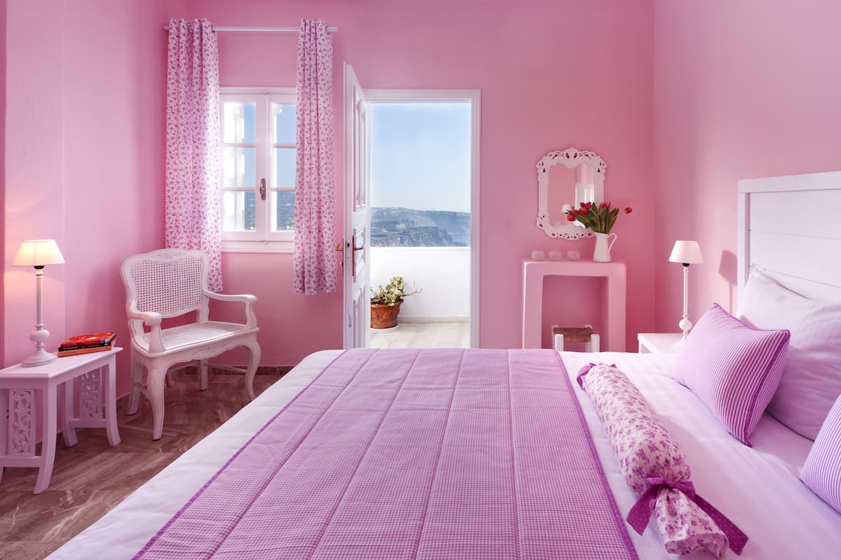 Et rosa værelse med seng, stol, sengeborde og udsigt gennem en åben dør.