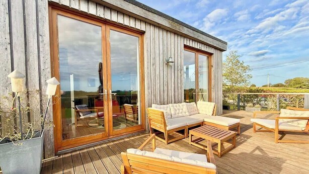 A wooden deck with outdoor furniture, large sliding glass doors, and a view of the landscape. - Grand Chapel with Views in Peaceful Devon Hills (Devon)