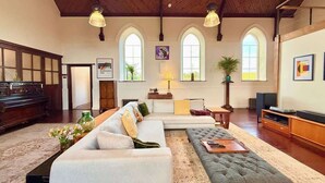 A spacious living room with a grand piano, a sectional sofa, a tufted ottoman, and a television. - Grand Chapel with Views in Peaceful Devon Hills (Devon)