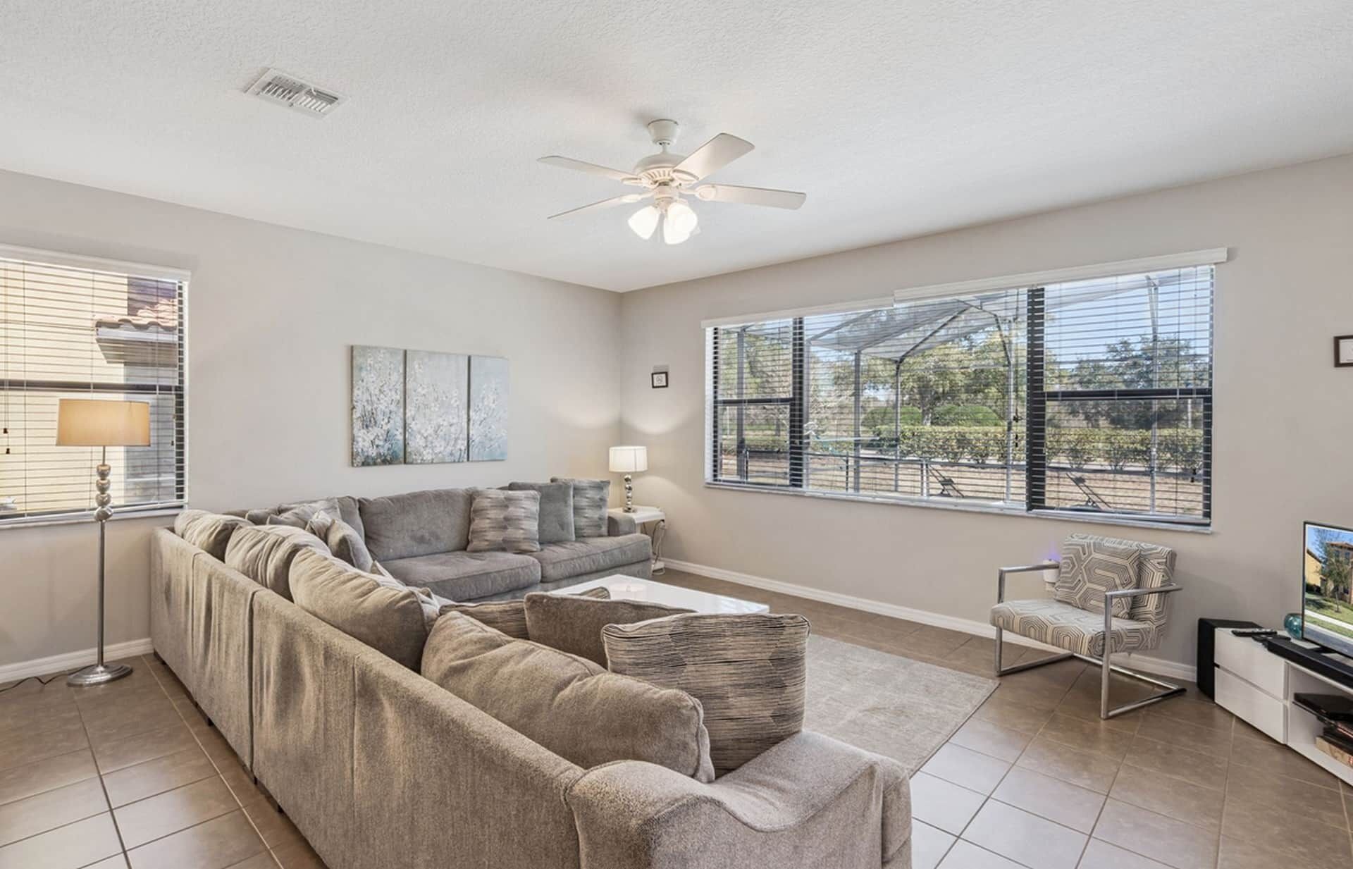 A living room with a sectional sofa, a small table, a chair, a ceiling fan, and large windows.