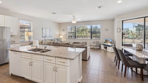Una cocina amplia con una isla central, un comedor con televisor y ventanas grandes que ofrecen vistas al exterior.