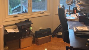 Une pièce comprenant un bureau, un ordinateur, une imprimante et une fenêtre donnant sur une maison et des arbres.