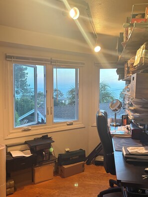 Ein Zimmer mit Schreibtisch, Computer, Drucker und einem Fenster mit Blick auf ein Haus und Bäume.
