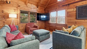 Ein Interieur einer Blockhütte mit einer Sofagarnitur, einem roten Sessel, einem Fernseher und einem Fenster mit Jalousien.