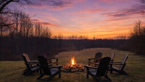 Ein Lagerfeuer mit vier Adirondack-Stühlen, die darum angeordnet sind, in einem grasbewachsen Feld mit Bäumen im Hintergrund.