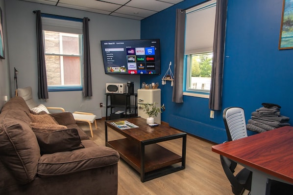 A living room with a brown couch, a wooden coffee table, a flat-screen TV mounted on the wall, and a window with curtains.