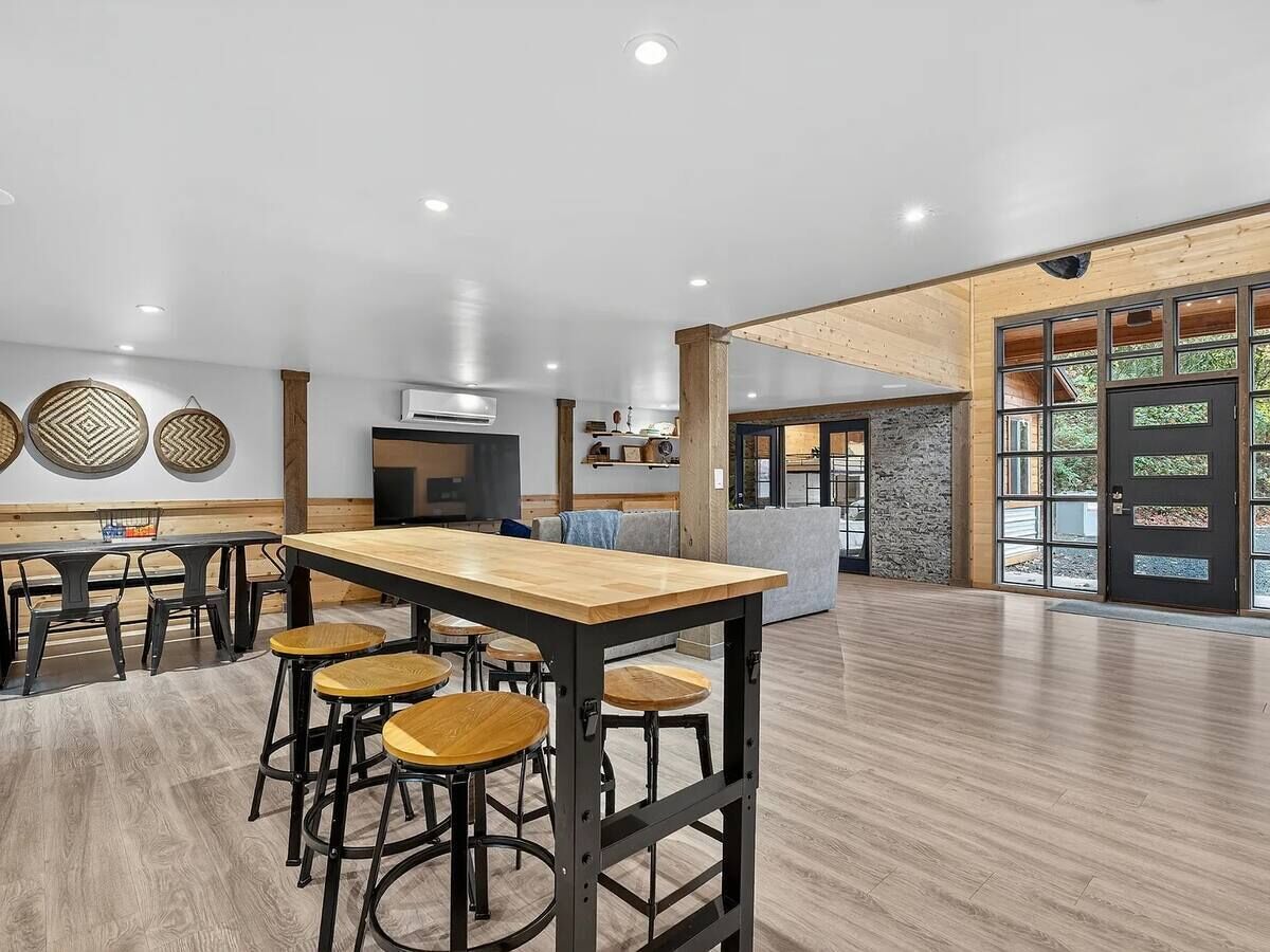 A spacious room with a long wooden table and bar stools, a kitchen area, and a large window with a view of greenery.