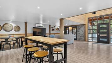 A spacious room with a long wooden table and bar stools, a kitchen area, and a large window with a view of greenery.