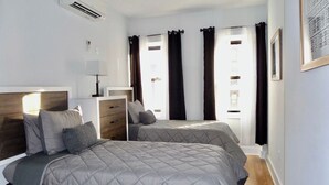 A bedroom with two beds, a bedside table with a lamp, a dresser, and a window with curtains.